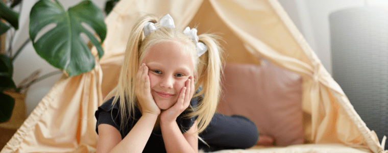 A little blond girl sitting in front of beige teepee