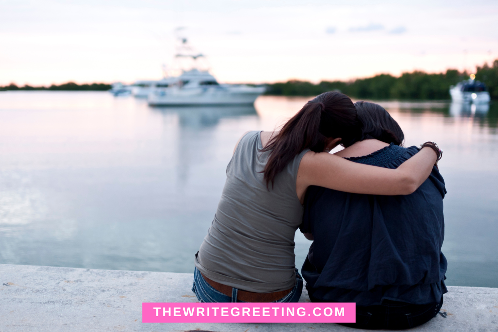 Two women sitting by water hugging